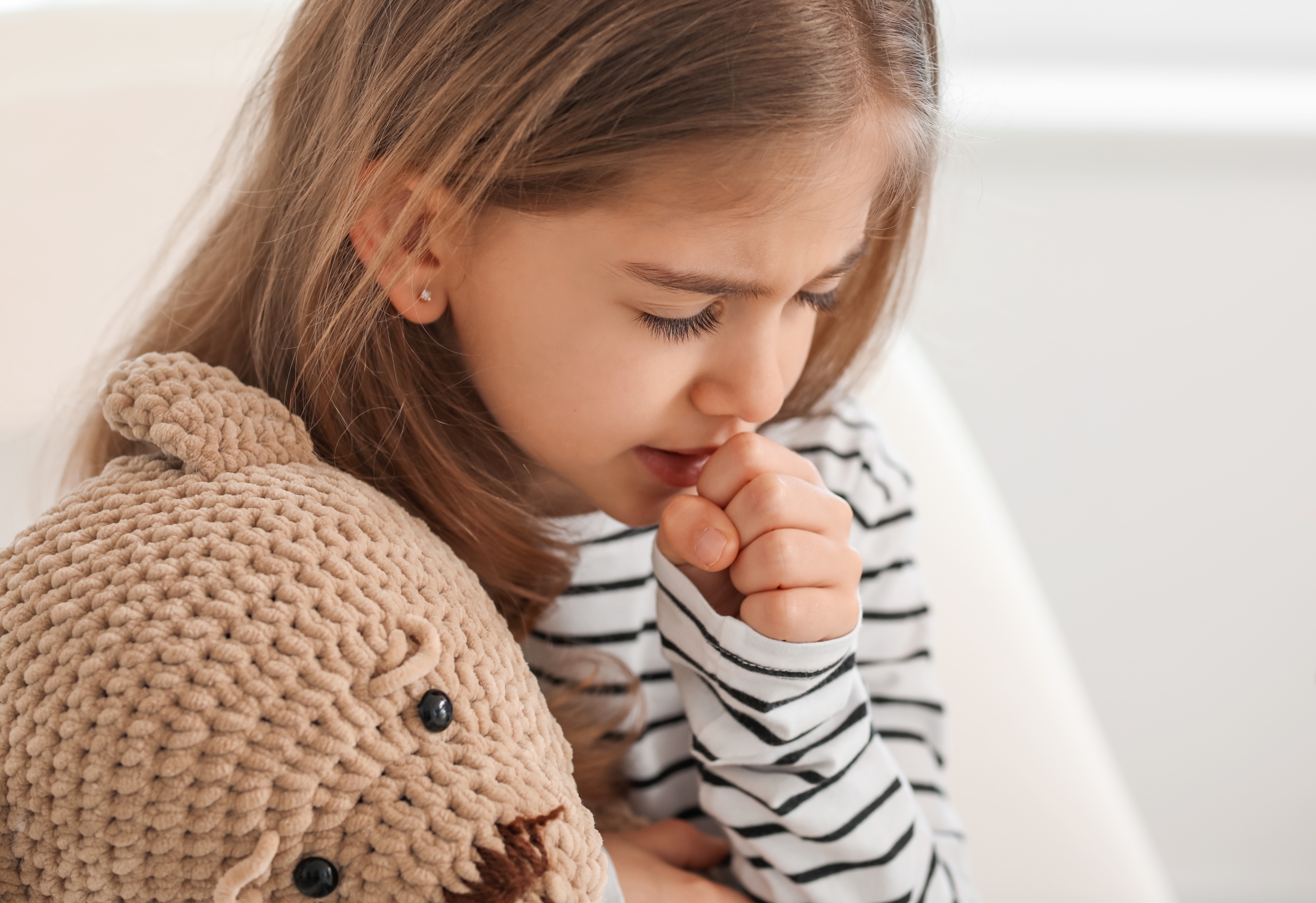 A little girl hugging her stuffed animal and coughing.
